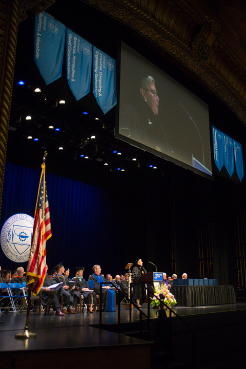 Below banners representing the college's six schools and a large screen, Penn College President Davie Jane Gilmour welcomes the commencement audience.