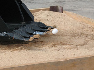 A "rodeo" participant delicately lifts an egg from the sand with a tablespoon taped to a backhoe's bucket tooth.