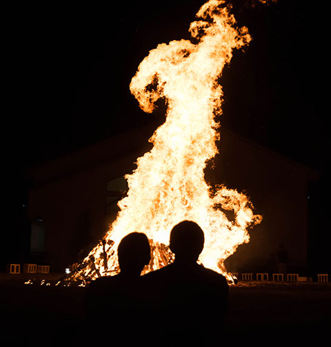 Flames lick the evening sky, silhouetting bonfire spectators.