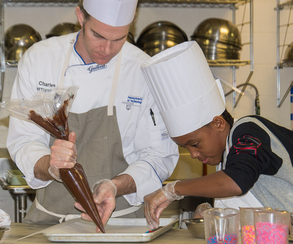 A middle school student adds M&Ms to rounds of melted chocolate piped by Chef Charles R. Niedermyer, instructor of baking and pastry arts/culinary arts, as they make knackerli.