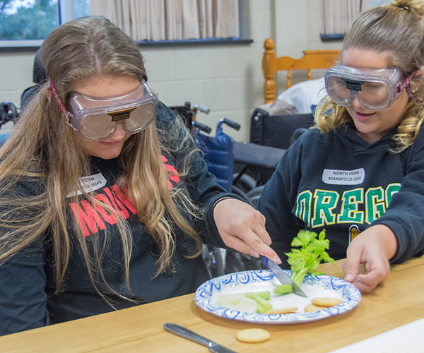A girl from North Penn-Mansfield High School enlists the help of her neighbor as she tries to cut celery and add peanut butter to crackers while wearing goggles that simulate a vision impairment and keeping her dominant hand behind her back. In the occupational therapy assistant program, such exercises help students to develop empathy for the clients they will serve.