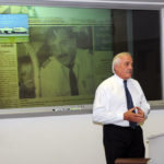 The speaker stands before a newspaper clipping, recounting one of his relief flights from Romania.
