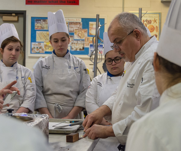Scourlas demonstrates a technique that involves dipping strips of acetate in melted chocolate to wrap pieces of chocolate gateau.