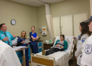During an Interdisciplinary Professional Education event at Penn College, students from several majors discuss a patient case, including what treatment each professional would provide and what information each would gather and share with professionals from other departments. The patient, in bed, is played by physician assistant student Danielle M. Klock, of Sunbury. Facilitating the discussion is Wayne E. Reich (left), director of nursing, bachelor degrees.