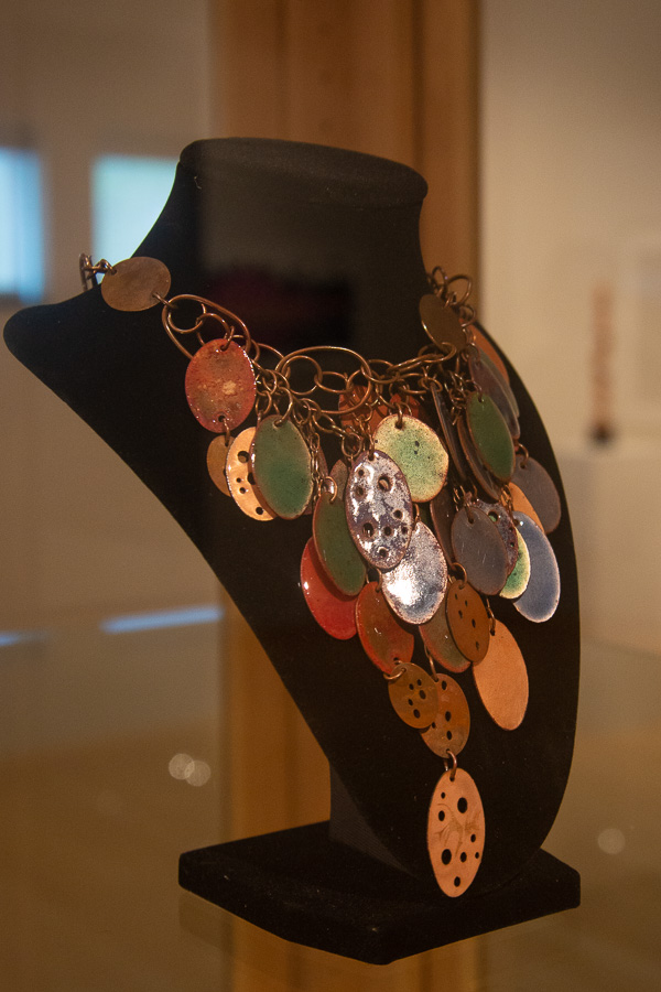 “Many Pennies,” a necklace of copper, pennies and vitreous enamels, shimmers in a glass display case.  