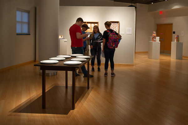 Students, visiting the exhibit for a class, review their notes. A table set with decorated porcelain plates – a work titled “Consumption” by Ian Thomas – is in the foreground.