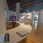 Students’ portfolios are offered for exploration on two curved tables along a gallery wall.