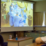 Outside the surgical technology lab’s mock operating room, PA students fulfilling the roles of emergency room and hospital staff members observe the work of their classmates.