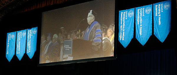 The overhead video feed, flanked by banners of the college's six academic schools, broadcasts the president's final remarks.