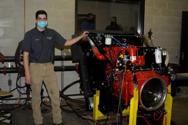 Michael J. Sormilic, an online power generation student from Southbury, Conn., who has spent much of the Fall 2020 semester learning the ins and outs of the equipment, stands alongside a Cummins crane engine donated to Penn College by Terex Corp. Behind him, in the control room of the dyno lab, are John D. Motto (left), diesel equipment technology instructor, and Chris Macdonald, assistant director of corporate relations.