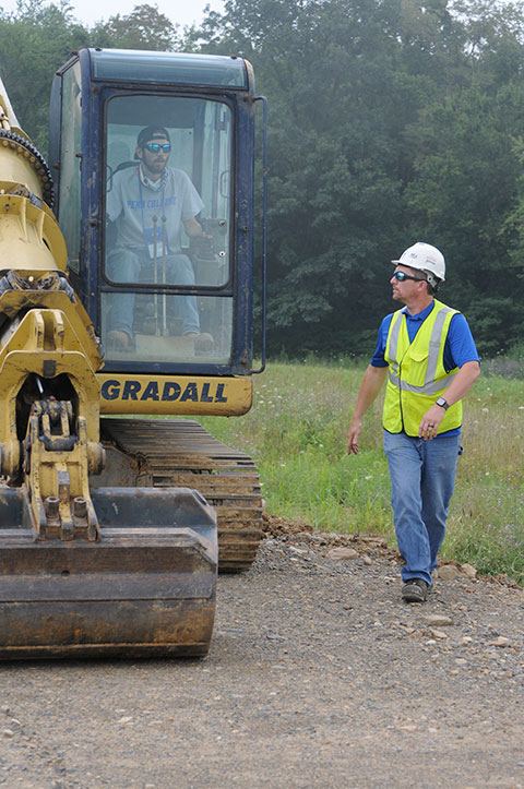 Diesel equipment technology instructor Chris S. Weaver provides walk-along guidance to a young man operating a crawler excavator.