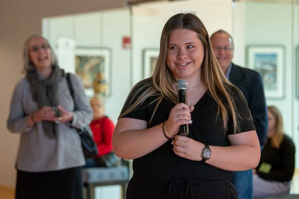Bookended by two of her faculty in the background – Kathryn M. Anderson (left) and Flynn (right) – Lexi “Alexa” C. Hamm, of Kutztown, offers humorous commentary. 
