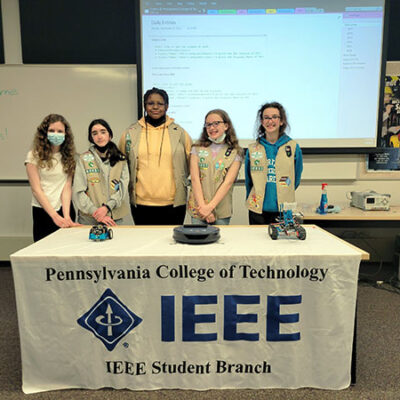 The five Cadettes pose in lab, amid robots and the IEEE chapter's banner.