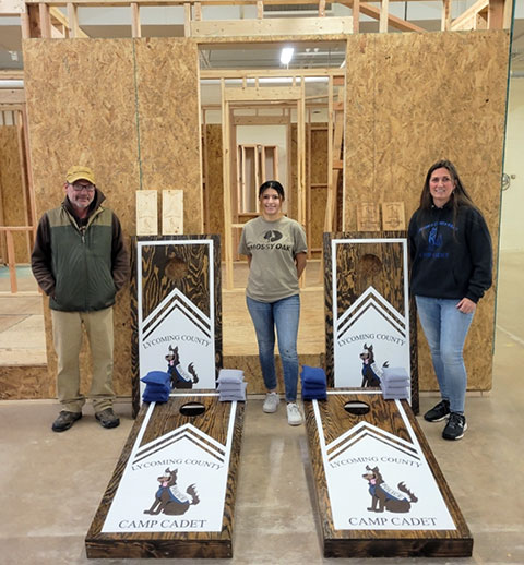 Cpl. Jennifer McMunn (right), representing the Pennsylvania State Police Office of Community Engagement, stops by the Carl Building Technologies Center on Friday to pick up student-crafted cornhole boards and award plaques for use at Camp Cadet. Also on hand are Barney A. Kahn IV, instructor of building construction technology and an adviser to the Penn College Construction Association, and PCCA President Lizeth V. Reyes-Becerra.