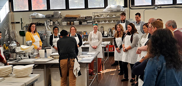 The group leans in, aproned and attentive, for its kitchen tutorial.