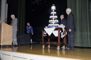 Marking her 10th year as Penn College president, Davie Jane Gilmour is surprised with a commemorative cake at Thursday's all-college meeting. In honor of the anniversary, the college's Board of Directors has commissioned artwork that will be placed on the third floor of the Student and Administrative Services Center when renovations are completed there. The art will be a custom piece designed by Gilmour's husband, Fred, (second from right) an alumnus and professor emeritus. 'Her modesty and integrity are matched by her dedication to Penn College,' said board Chairman Robert E. Dunham (at podium) in announcing the tribute. 'I know of no one more dedicated to teamwork, to shared vision and to sharing the credit, whenever credit is due.' Joining in the surprise was Gene Yaw, college solicitor.