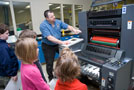 Jody L. Fausey, pressroom production specialist (with Raymond J. Fischer, manager of document services, through the glass) demonstrates the Heidelberg Speedmaster offset press