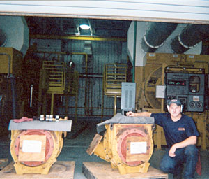 Matthew J. Strine kneels next to two CAT G3616 turbochargers. Sixteen-cylinder engines, powered by landfill gas, are visible in the background.