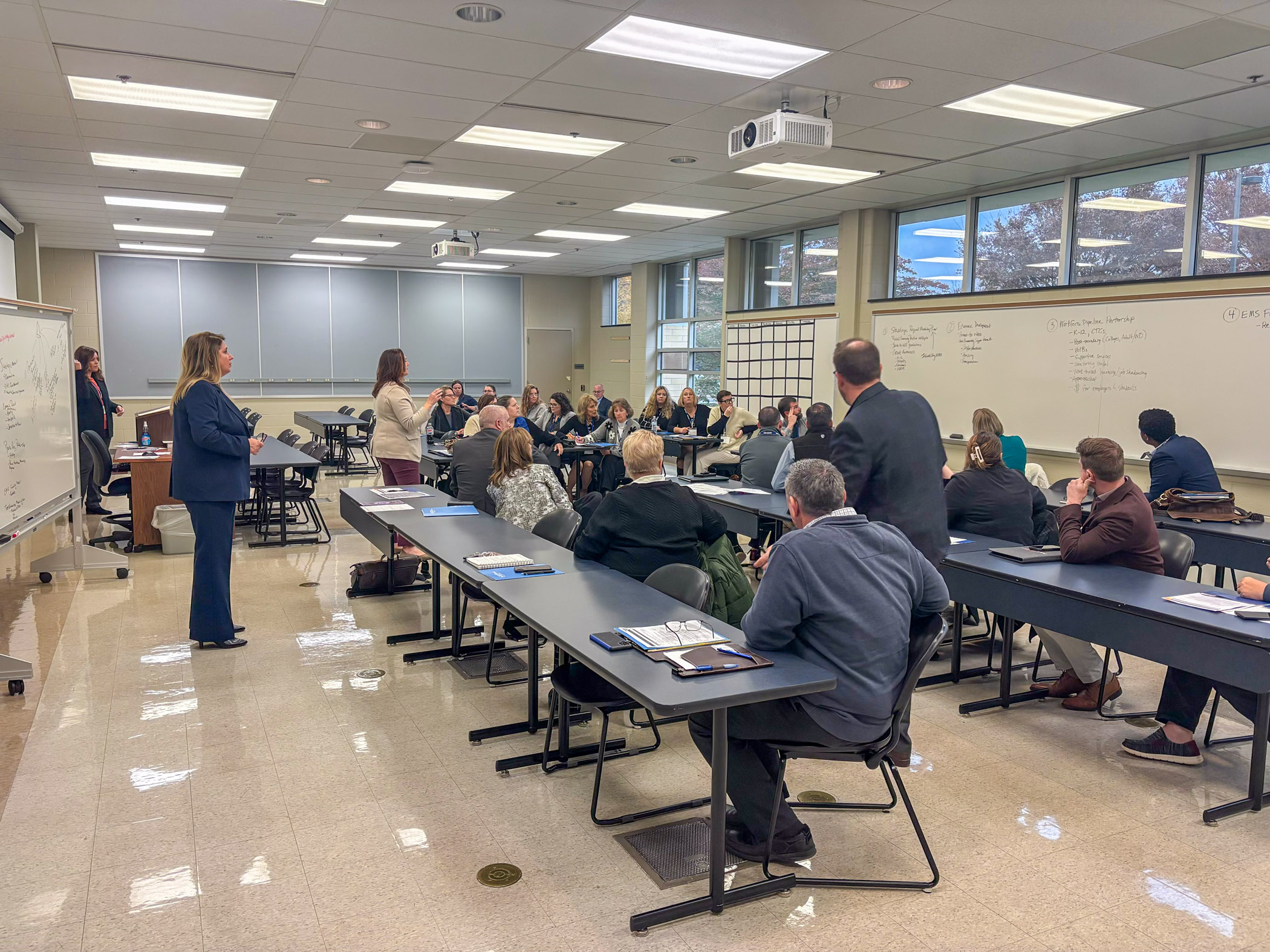 A classroom full of adults in business attire look at a whiteboard.