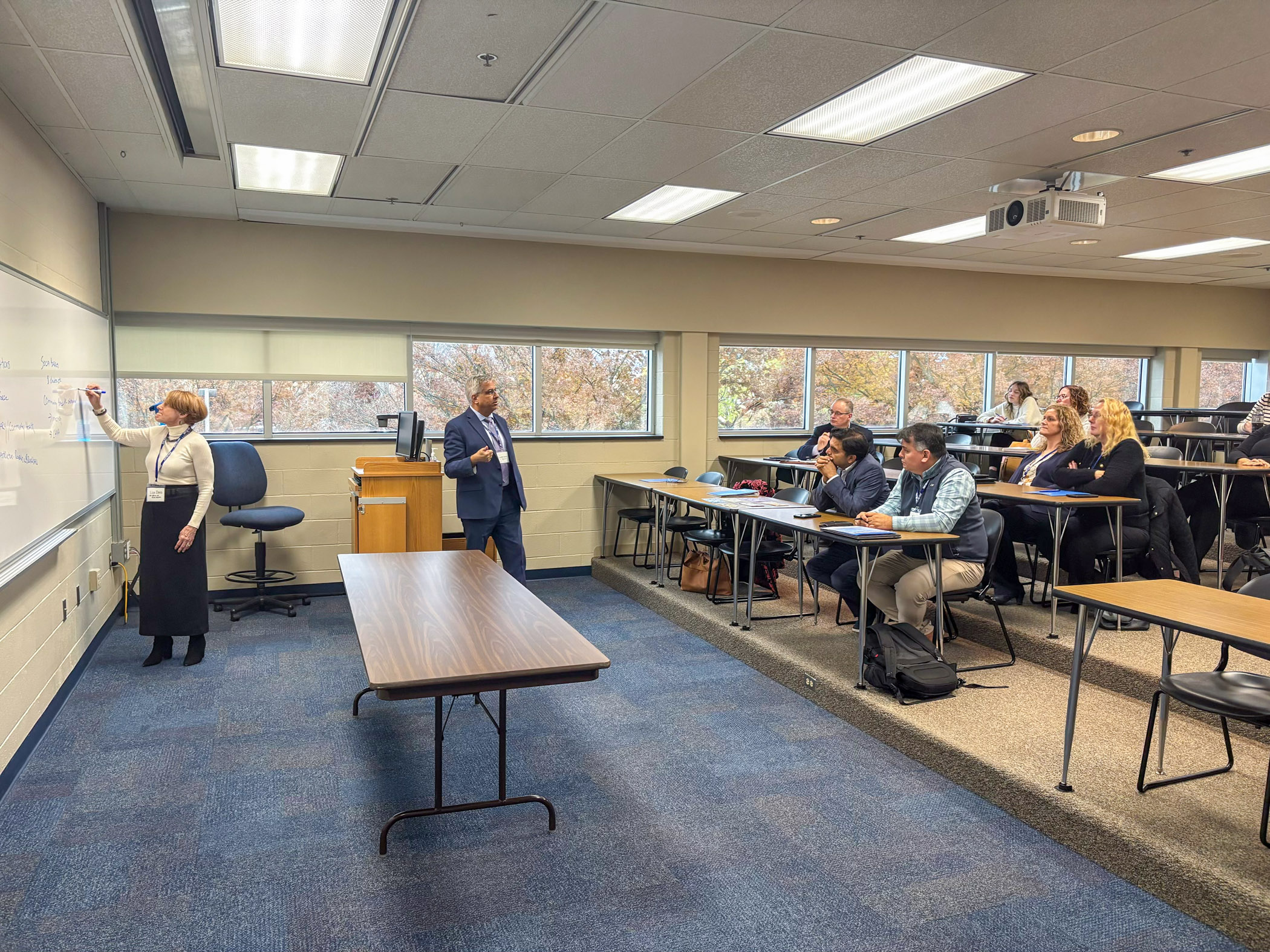 A classroom full of adults in business attire. A woman writes on a whiteboard while a man lectures.