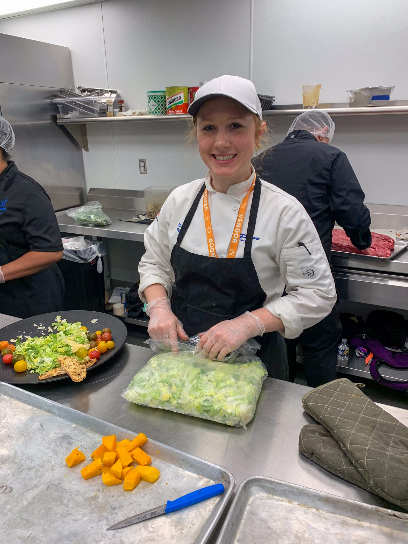 In a commercial kitchen, a student wears a chef's coat and apron and opens a bag of lettuce.