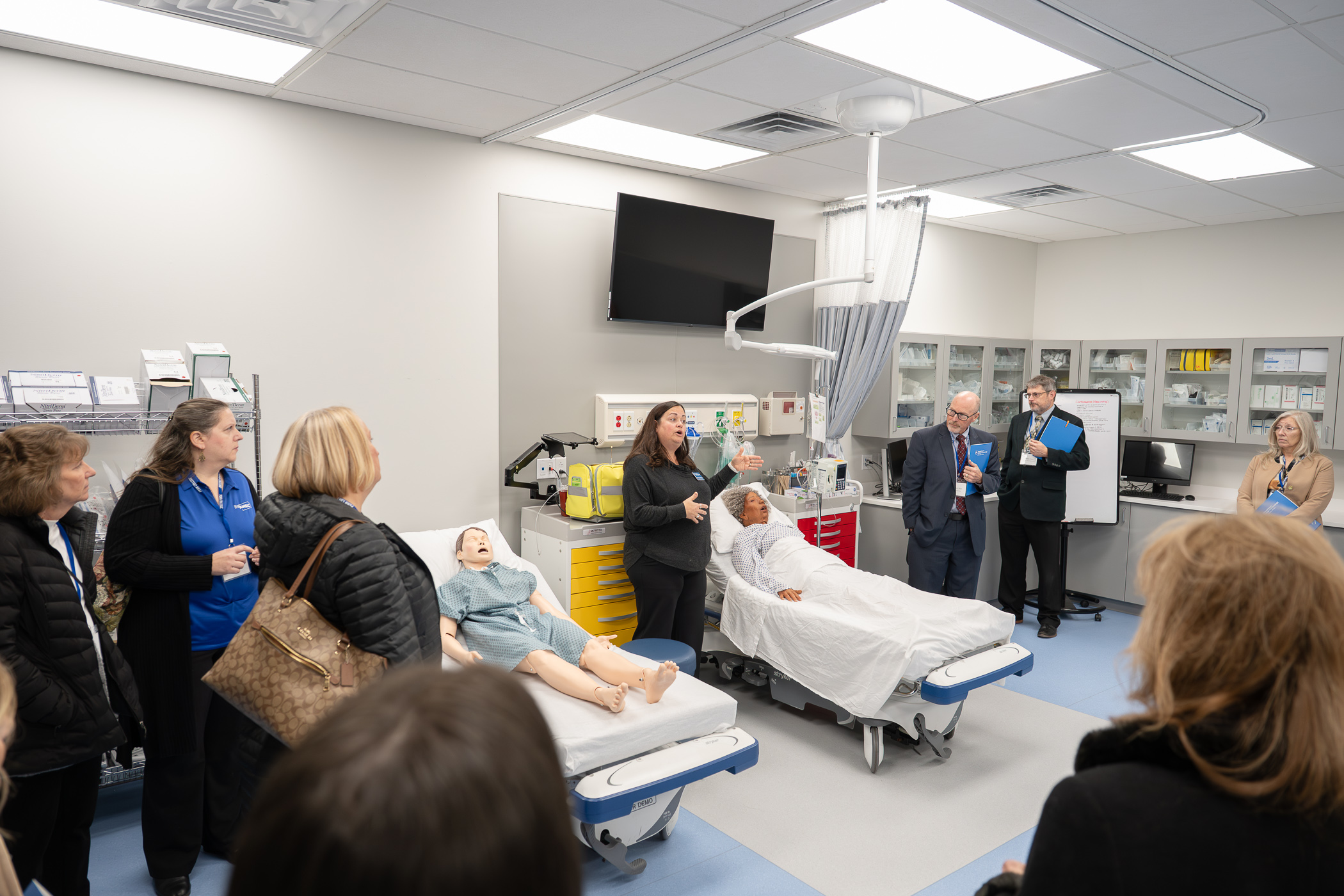 Melissa Sedor speaks to a group of people in business attire while standing next to hospital beds occupied by manechins in the Penn College Physician Assistant Center.