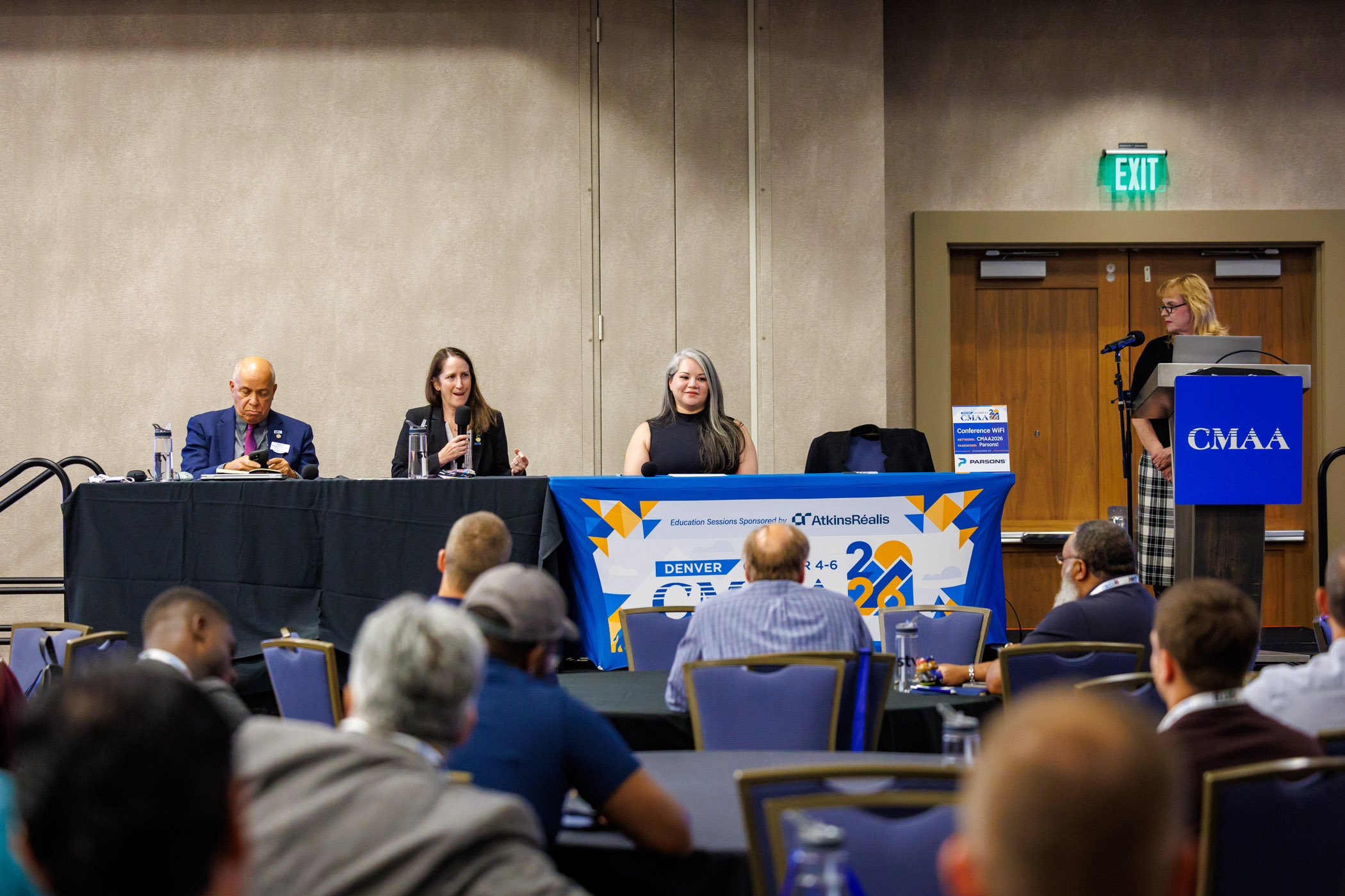 Audience members listen as a conference panelist speaks into a handheld microphone from a table on stage, while another presenter, Ellyn Lester, stands at a podium during a CMAA conference session.