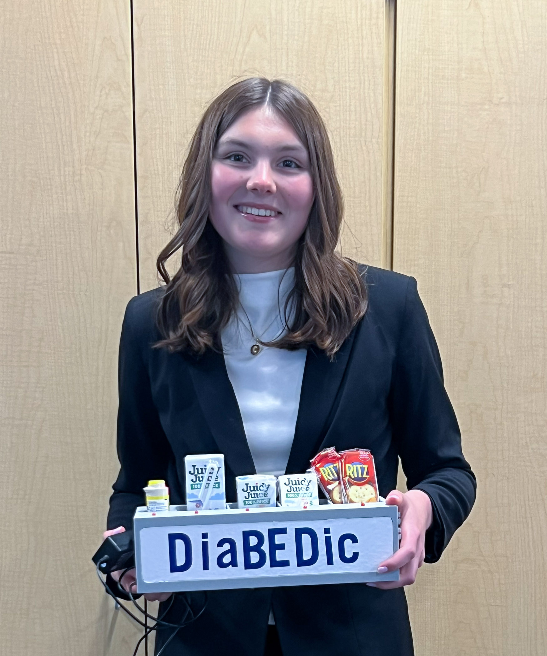 A high school student in a business suit holds a box that says "DiaBEDic" and has compartments with lights that hold a variety of snacks and juices.