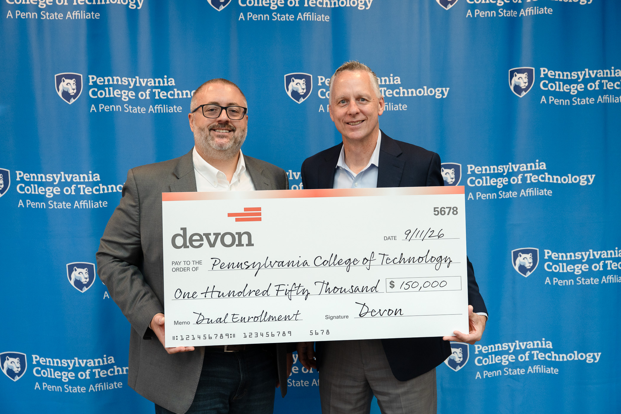 Two men in business attire hold an oversized check. They are standing in front of a step-and-repeat backdrop that has Pennsylvania College of Technology logos.