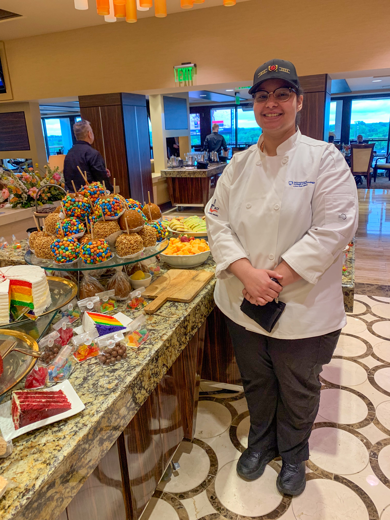 Student in chef uniform stands near a large display of dessert items.