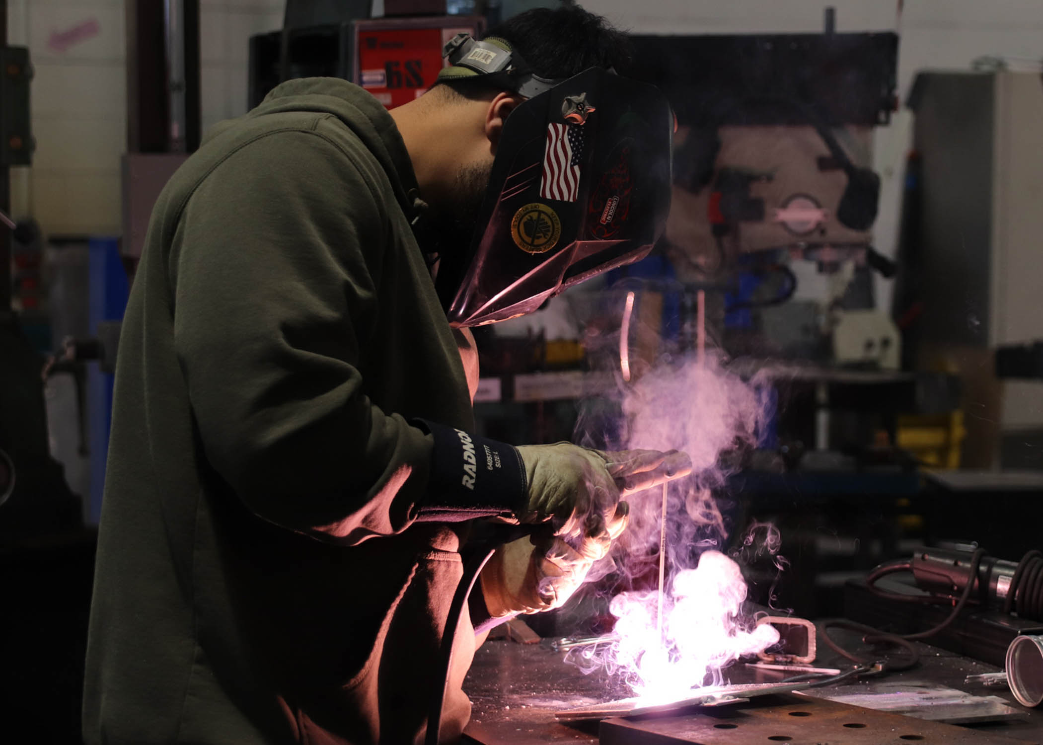 A student in a welding helmet and protective gloves welds a piece, creating light and smoke.