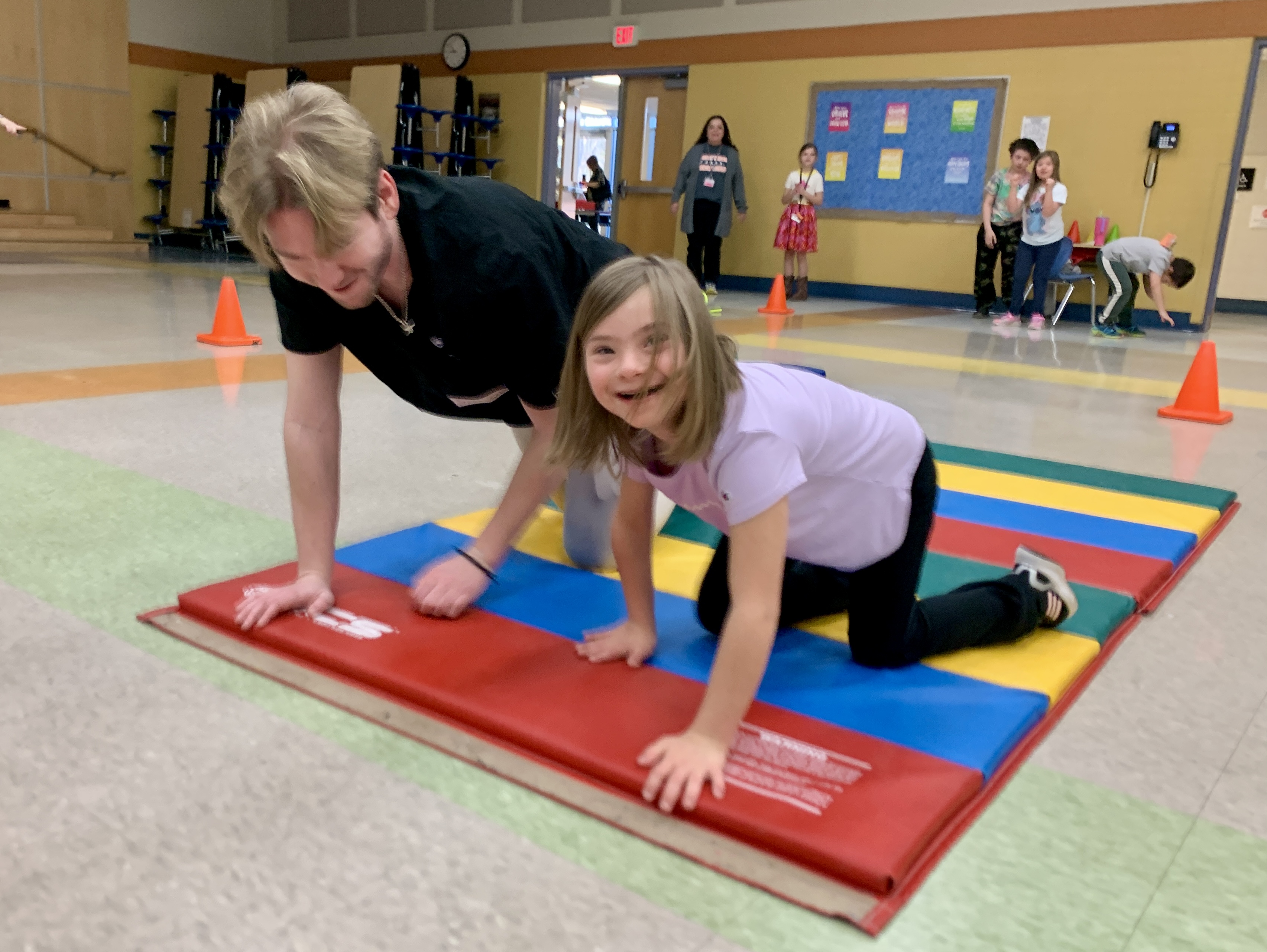 A college student and elementary school student crawl across a gym mat.