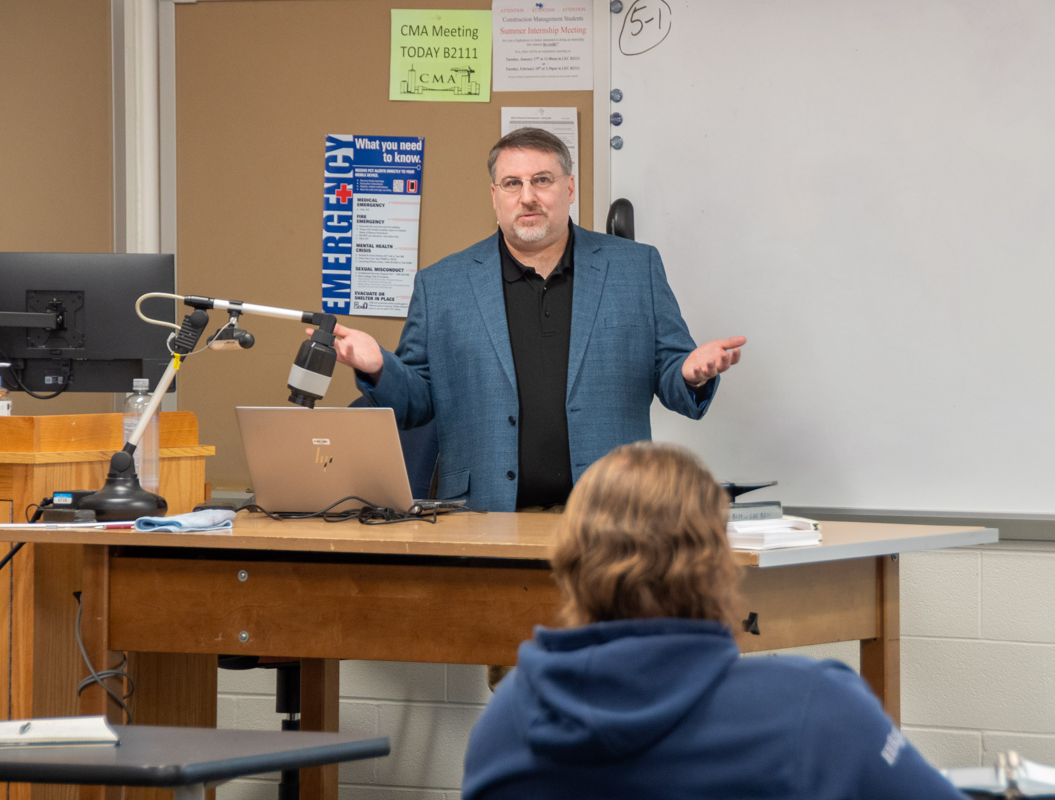 A person gestures while speaking in front of a construction management classroom.