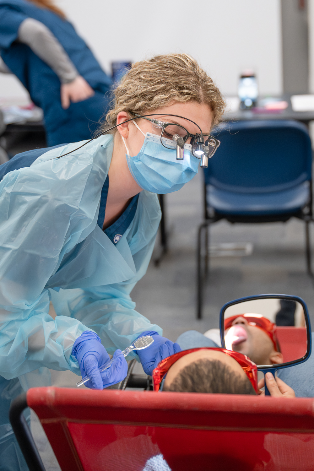 A dental hygiene student in medical protective gear wears loupes and holds a dental mirror while talking with a client who is laying back in a dental chair.