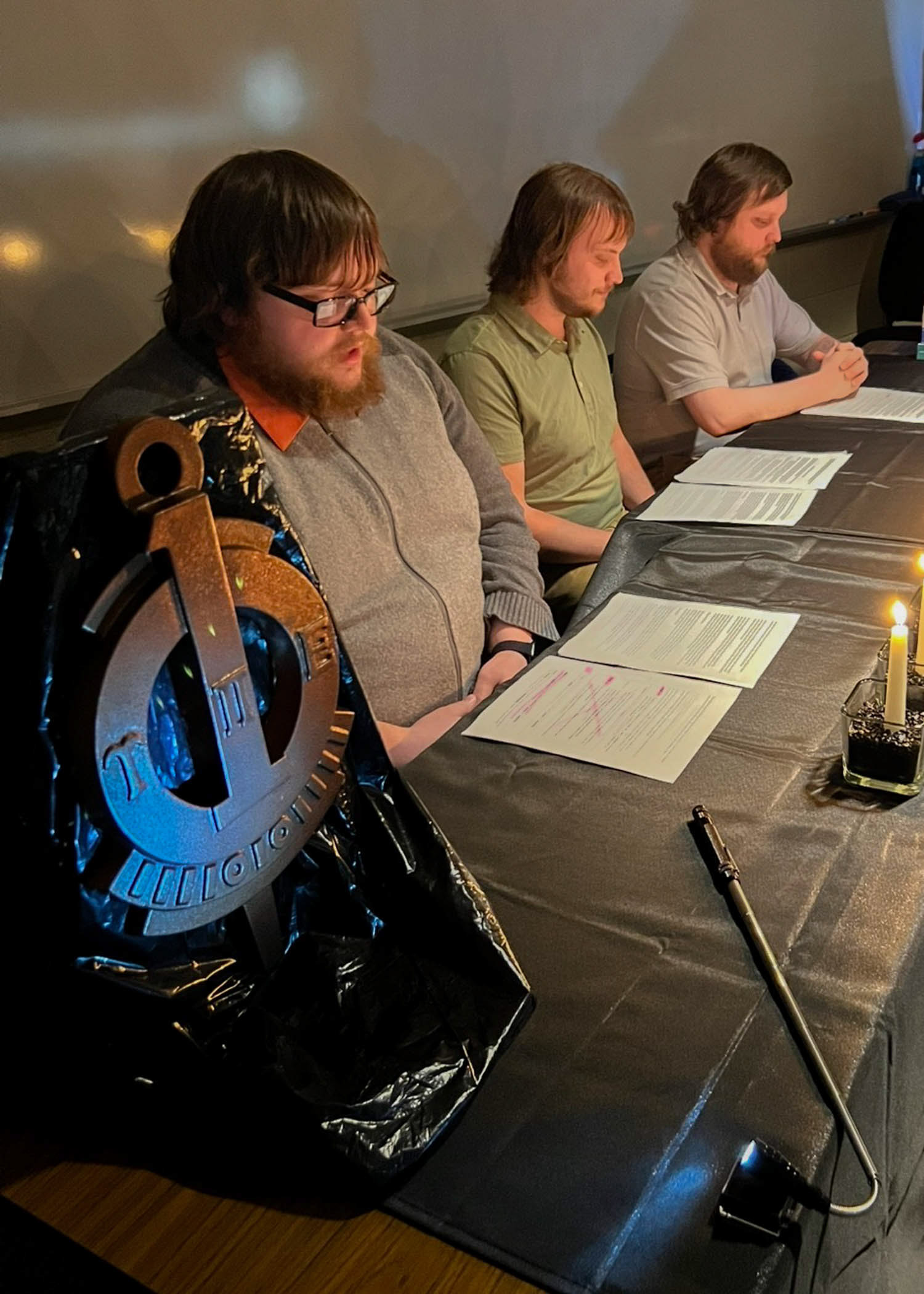 Students sit behind a candlelit table. The table holds a large UPE key.