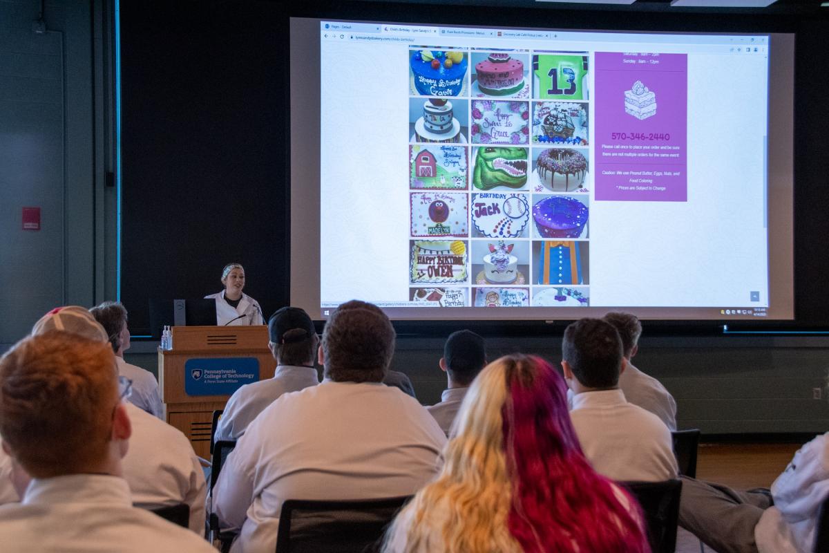 Summa talks about life as a bakery owner with students in Facilities Planning. (Some of her bakery’s creative cake designs, shown off on the Lynn Sandy’s Bakery website, shine on-screen.)