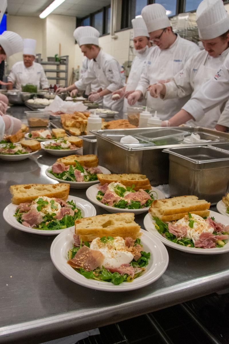 A salad of arugula, shaved asparagus and radish – topped with burrata cheese, prosciutto, hazelnuts and herb focaccia – awaits pickup by dining room staff as students continue the plating process.