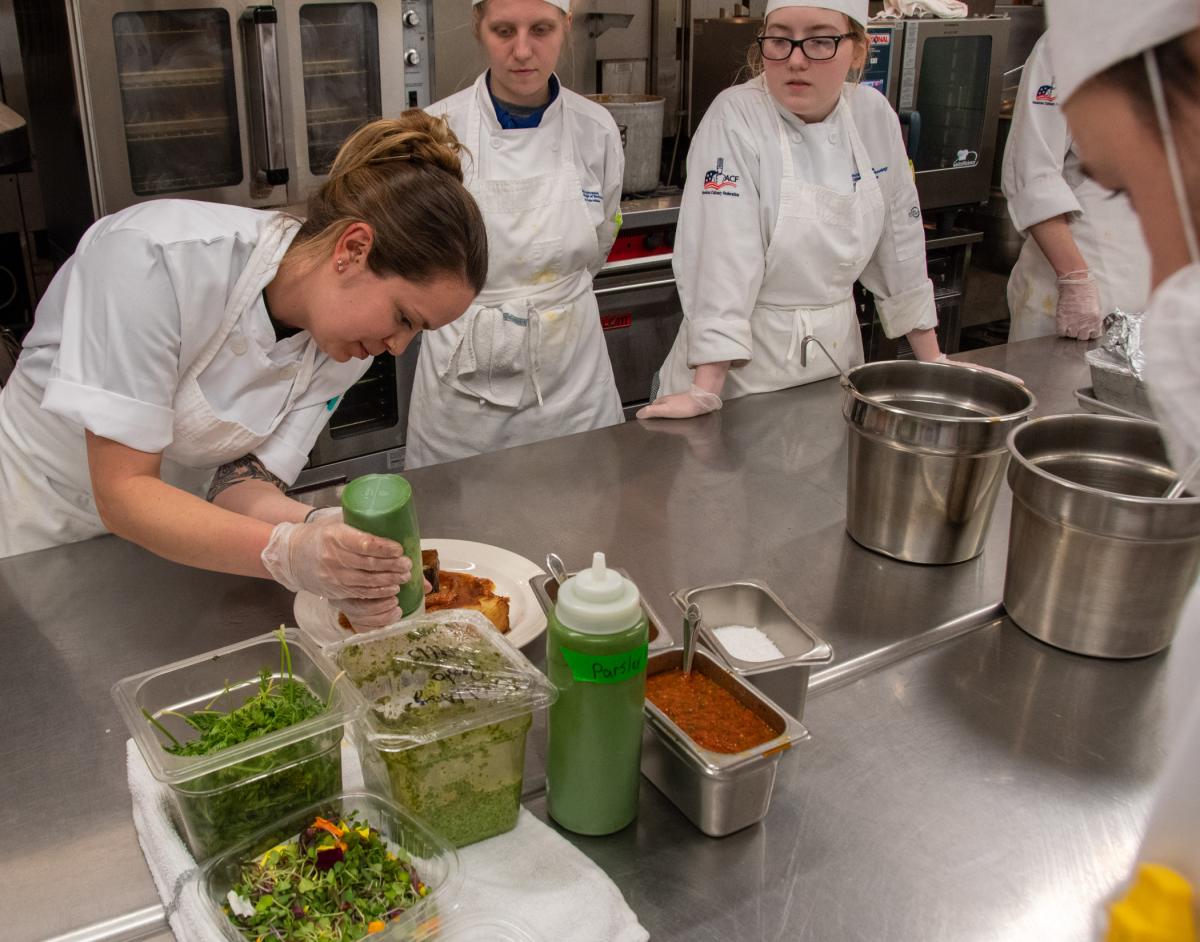 Demonstrating her main course’s plating scheme, Wisneski adds pureed parsley to a plate. Looking on are Walters (center) and Audrey N. Grello, a culinary arts technology student from State College.