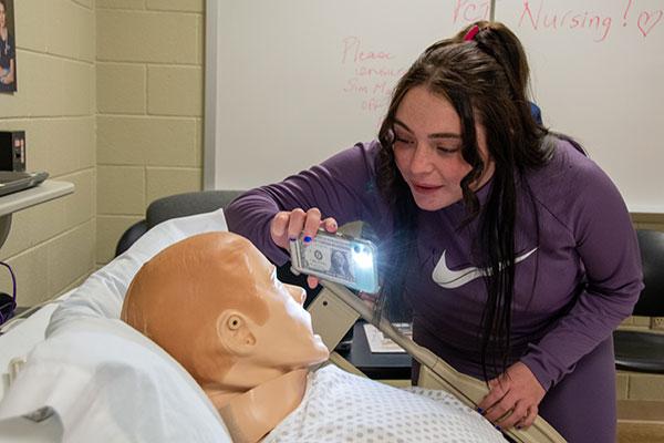 Future nursing student Kayla Mulvaney uses her phone’s flashlight to test the dilation of SimMan’s pupils in the college’s nursing facilities.