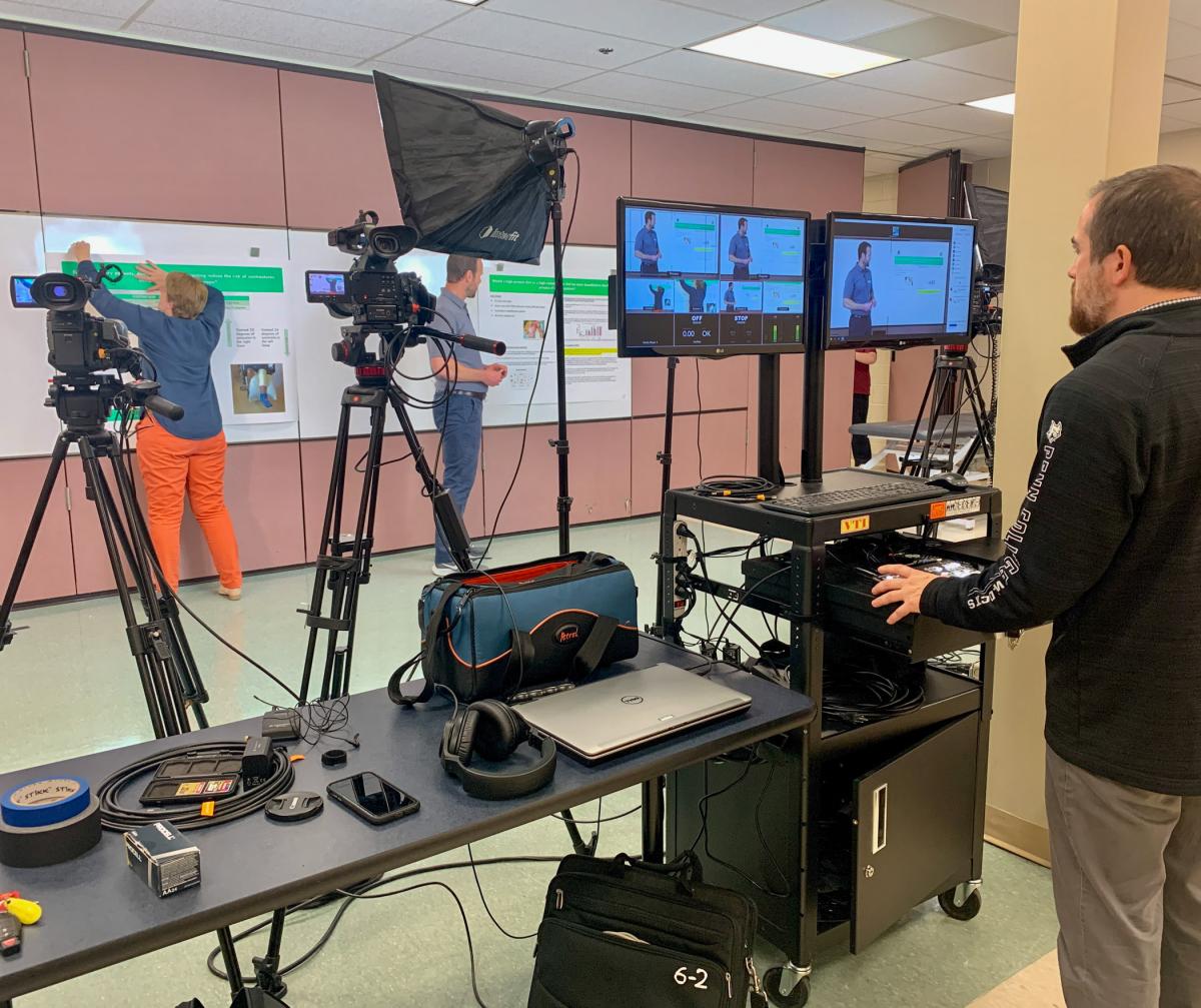 A glimpse behind the scenes: Tilburghelps to hang an off-camera poster while Moore presents on-camera, with Miller at the podium controls.