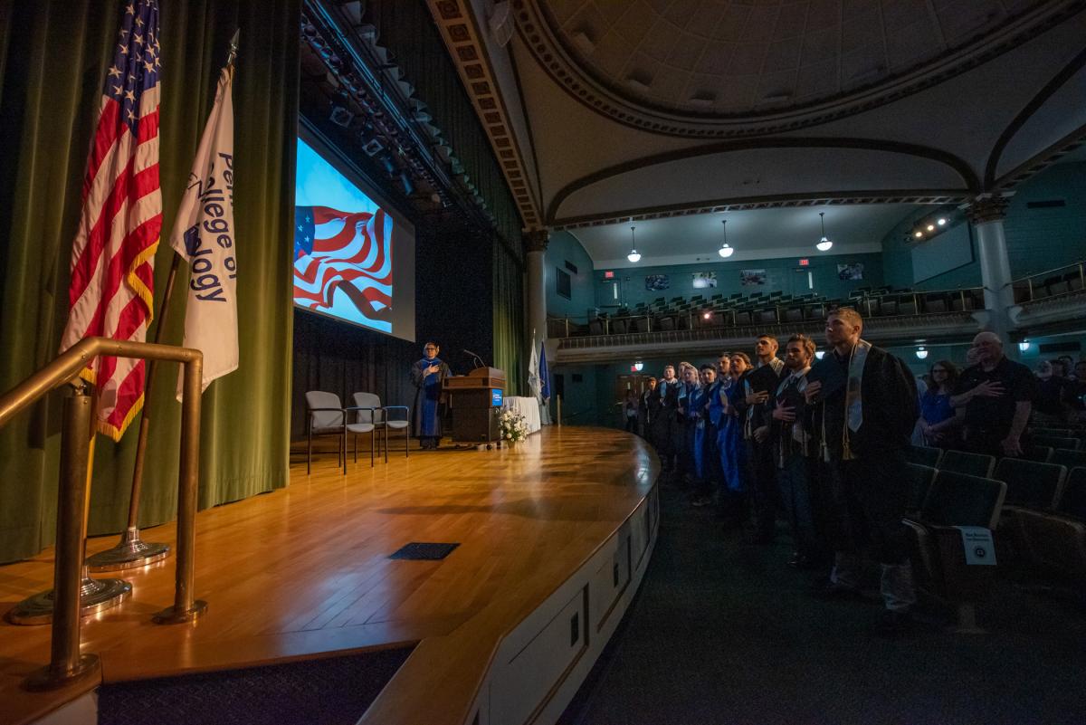 The crowd rises for the national anthem, a familiar part of graduations and ballgames, backed by projected photos of the large flag at the campus entrance.