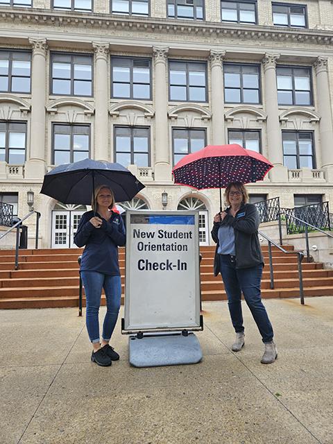 Warding off a Thursday morning drizzle with umbrellas and sunshiney smiles are Heather M. Shuey (left), senior director of employee success, and Sue A. Mahaffey, coordinator of enrollment management operations.