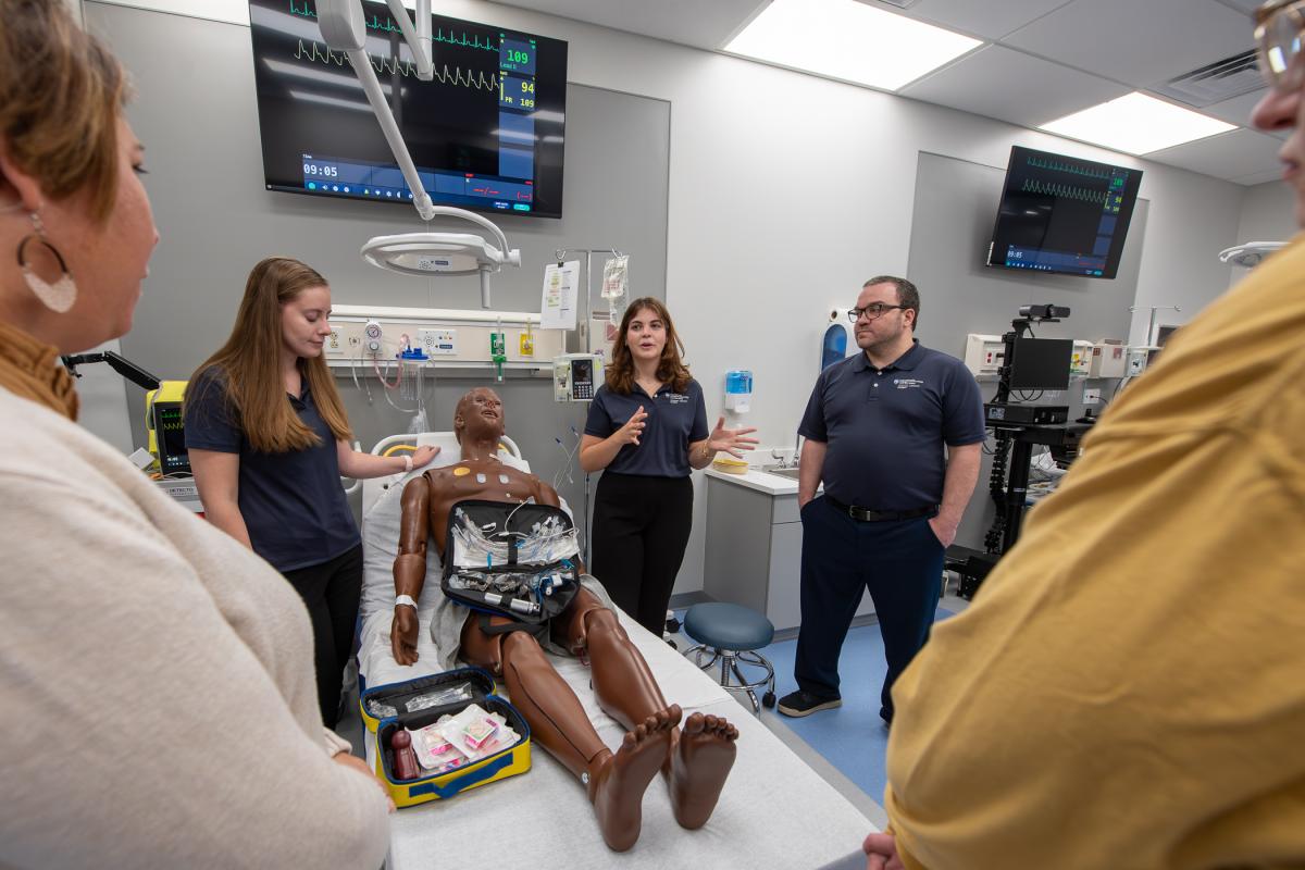 In the Emergency Medicine Lab of the college's Physician Assistant Center, students (in blue shirts) engage guests during an open house unveiling the center’s extensive renovations. From left, are: Sarah L. Fetterolf, of Millmont; Erin T. Braxton, of Berwyn; and Phillip Silvagni, of Williamsport. The manikin is HAL, an advanced, multipurpose patient simulator. 