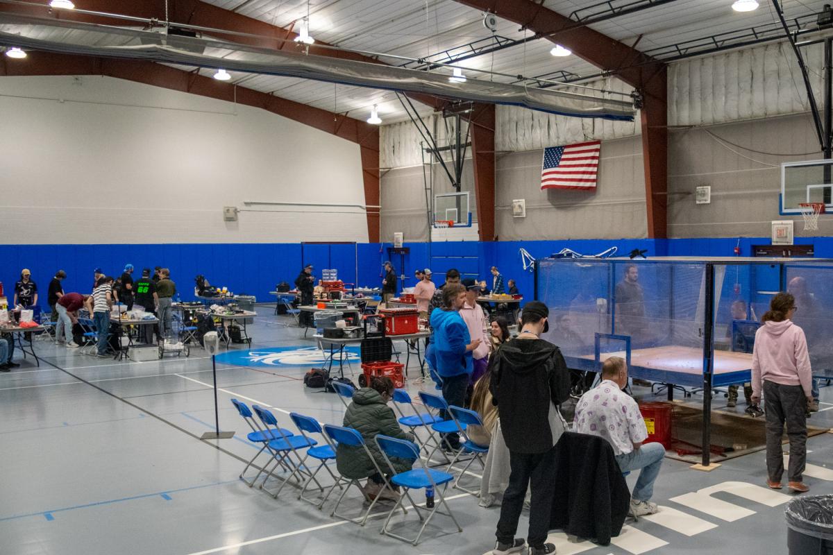 Competitors and spectators occupy the Field House, moving between prep/repair tables and the main attraction.