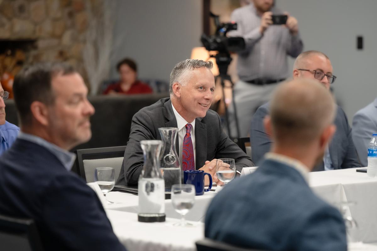 President Michael J. Reed smiles while sitting at a conference table with other people in business attire in the Thompson Professional Development Center.