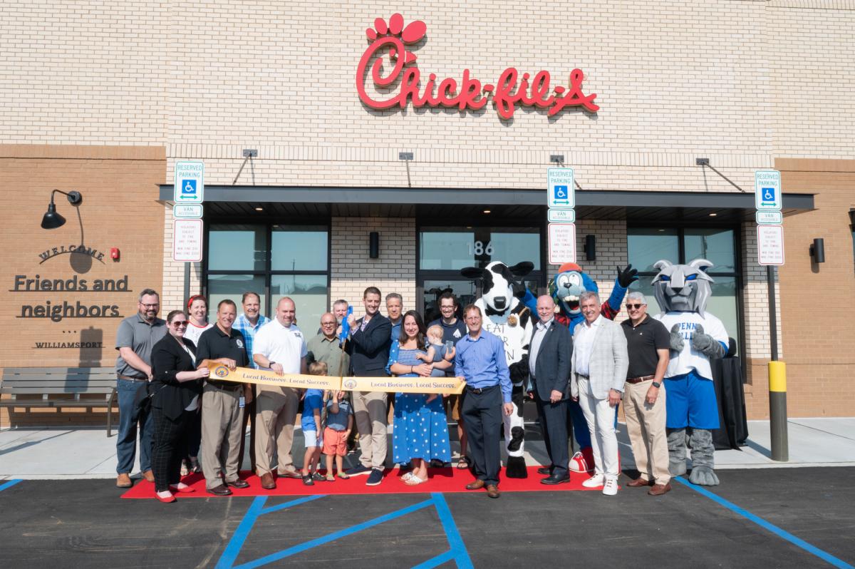 People, including three in mascot costumes, stand behind a ribbon at the entrance of a Chick-fil-A restaurant. One person is holding a giant pair of scissors up to the ribbon.