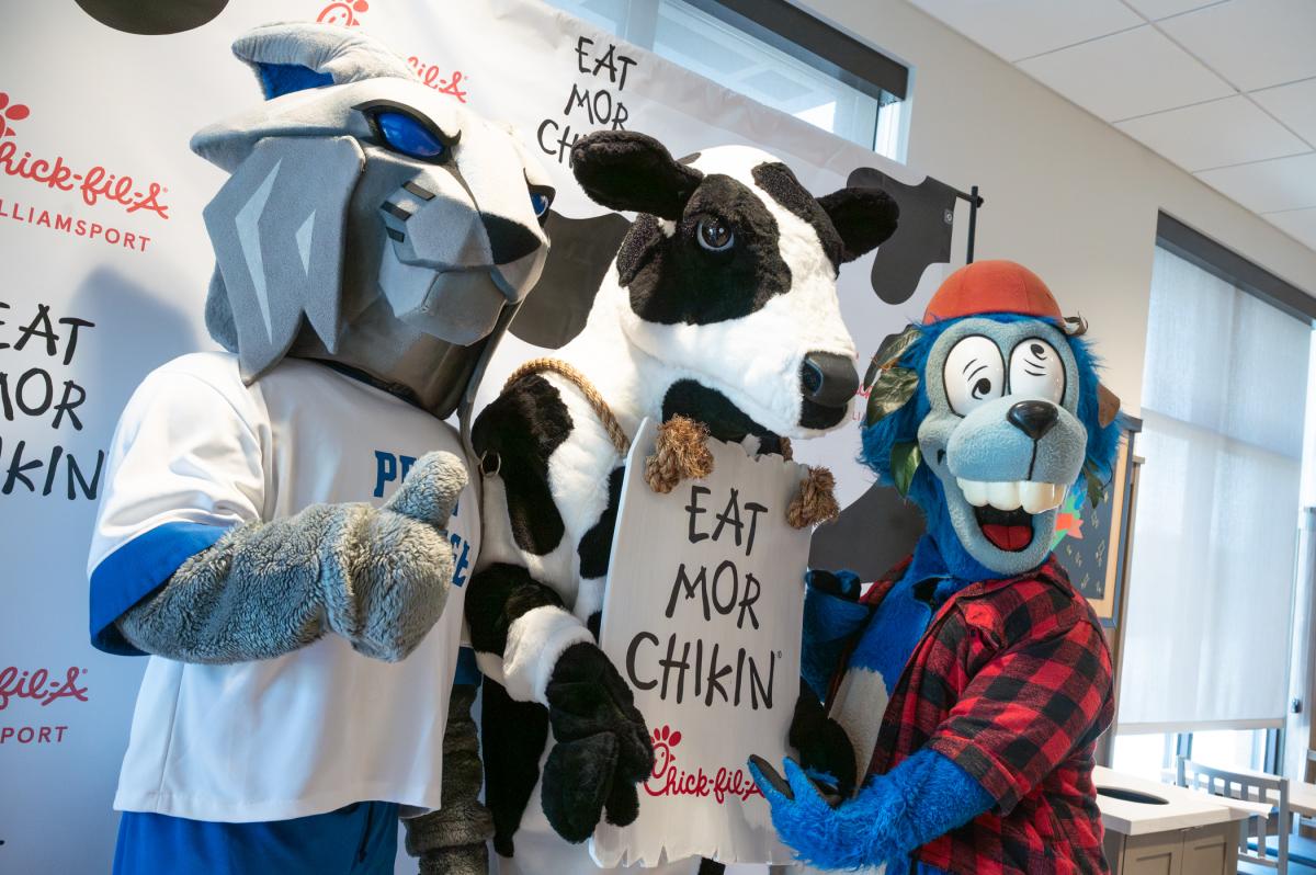 Three mascots: A wildcat giving a high five, a holstein cow holding a sign that says "Eat Mor Chikin," and a flurry blue character in a red buffalo-plaid shirt, pose together. 