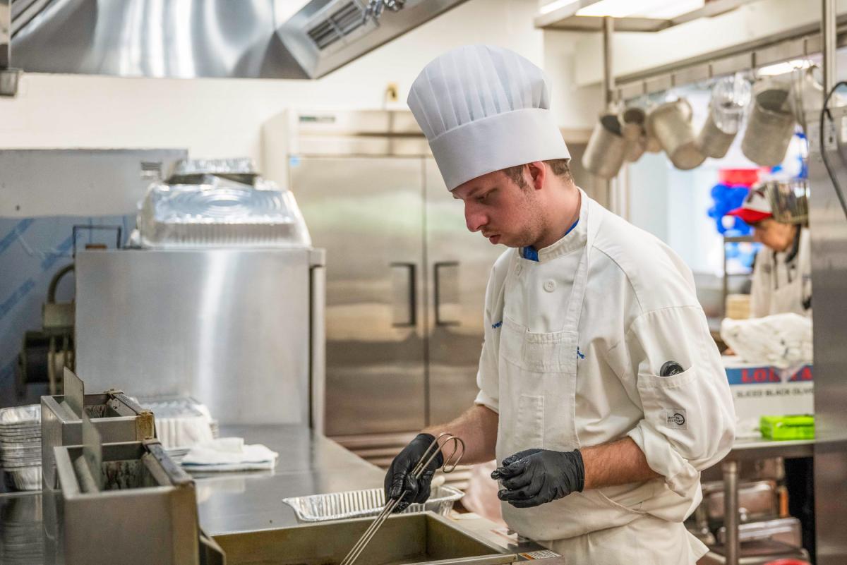 A young man in chef's uniform holds the handle of a fryer basket in a commercial kitchen.