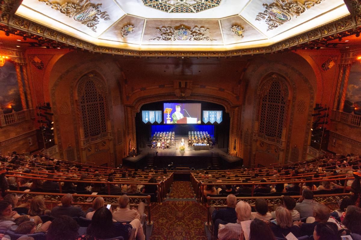 Interior of the Journey Bank Community Arts Center, showing ornate lighting fixtures above, lighted stage at front with a student in cap and gown speaking at a podium, and auditorium seats filled with audience members.