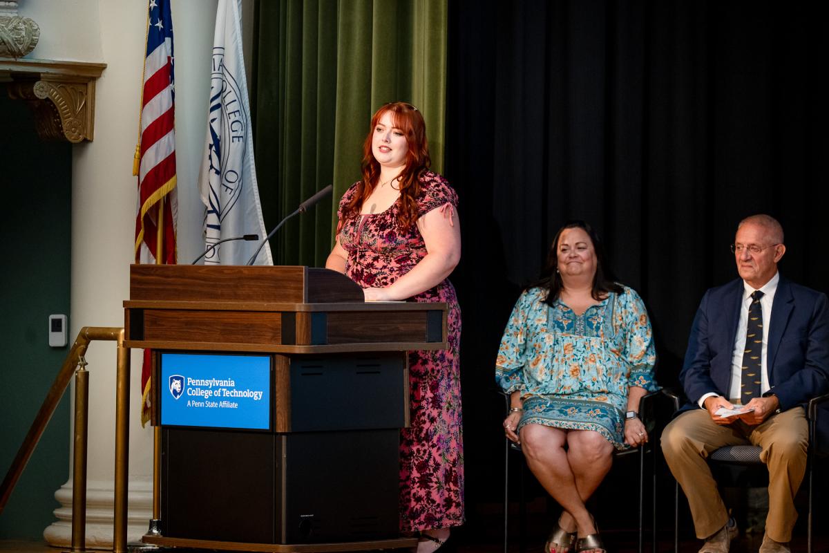 A woman in a dress stands behind the podium on the stage of Penn College's Klump Academic Center Auditorium.
